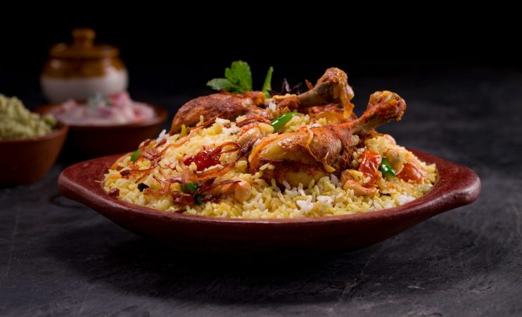 Chicken dhum biriyani using jeera rice and spices arranged in   an earthen ware with raitha and lemon pickle on grey background.